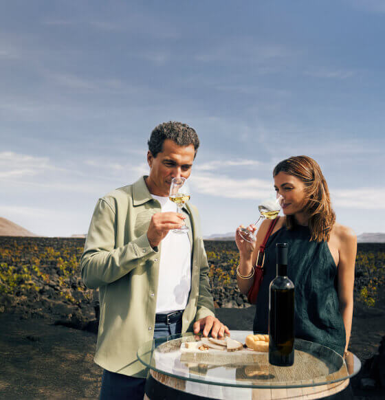 Couple en train de déguster du vin blanc sur une table en verre avec une bouteille et des produits locaux au milieu d’un paysage volcanique.