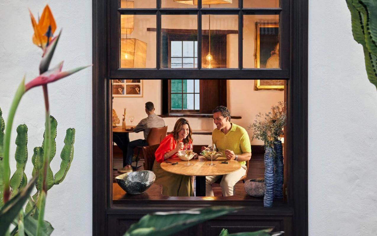 Vue à travers la fenêtre d’un couple en train de manger dans un restaurant chaleureux, avec une décoration traditionnelle et des détails naturels.