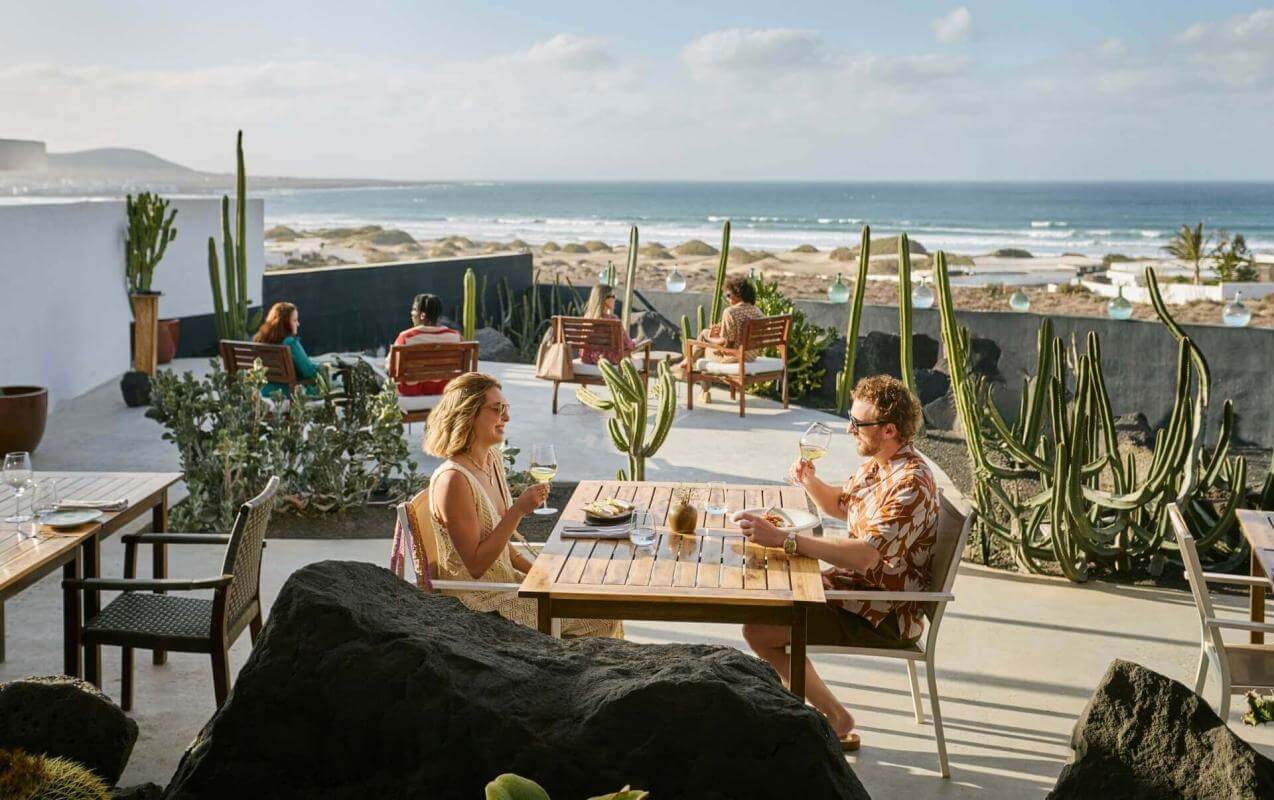 Personnes en train de partager un repas sur une terrasse avec vue sur la mer, entourée de cactus et avec un mobilier au design contemporain.