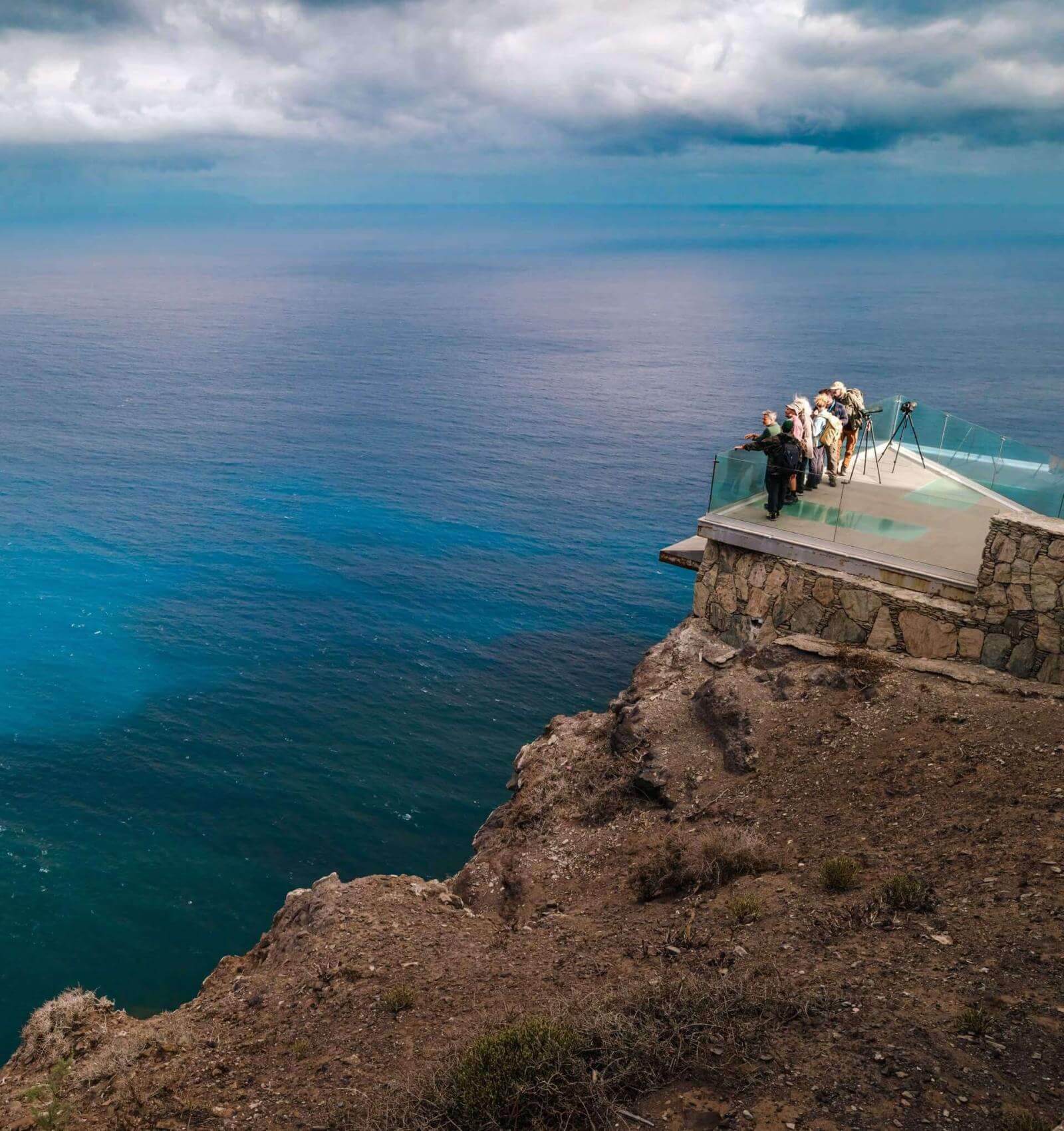 Personnes observant l’océan Atlantique depuis un belvédère en verre sur une falaise, sous un ciel nuageux.