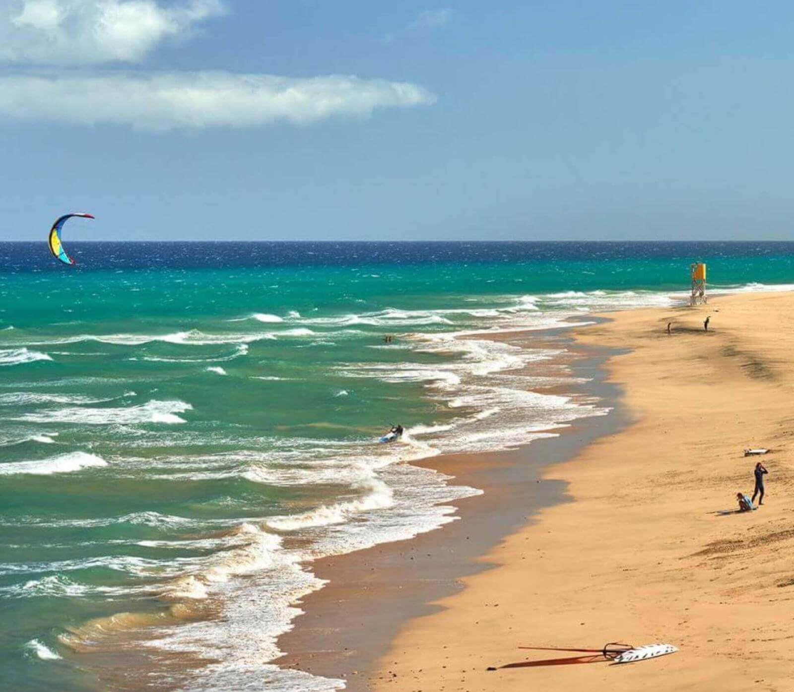 Plage de sable doré avec des vagues et des personnes en train de pratiquer des sports nautiques, comme le kitesurf.