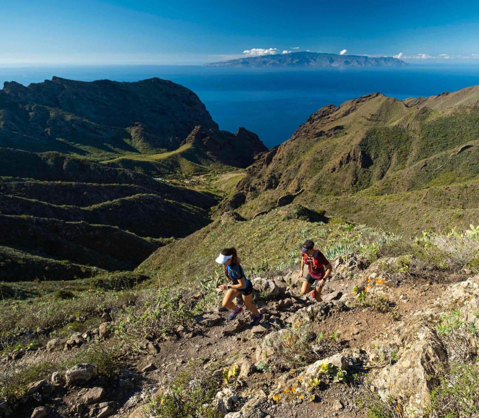 Deux personnes en train de pratiquer l’ultra-trail sur un sentier de montagne avec vue sur l’océan et une autre île à l’horizon.