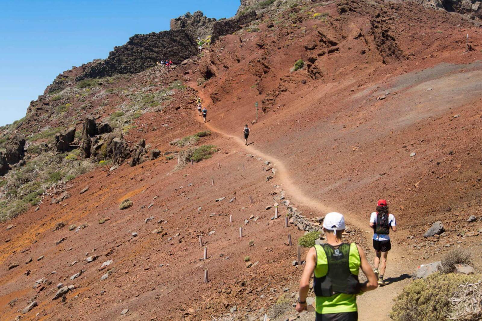 Coureurs en train de traverser un terrain volcanique rouge sur un sentier étroit et sous un ciel dégagé.
