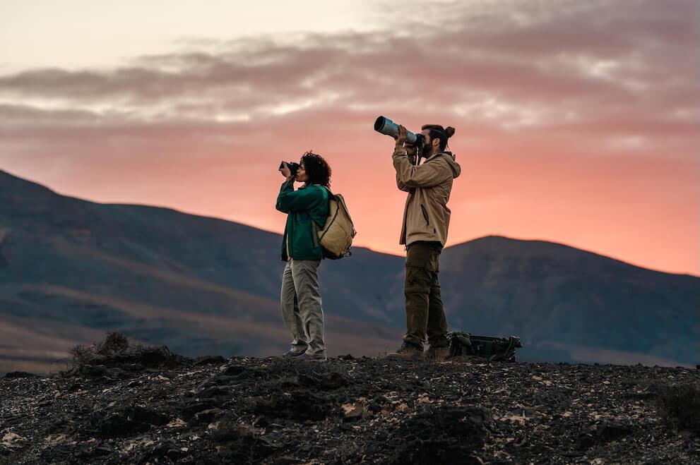 Deux personnes observant les oiseaux avec jumelles et téléobjectif dans un paysage volcanique à l’aube.