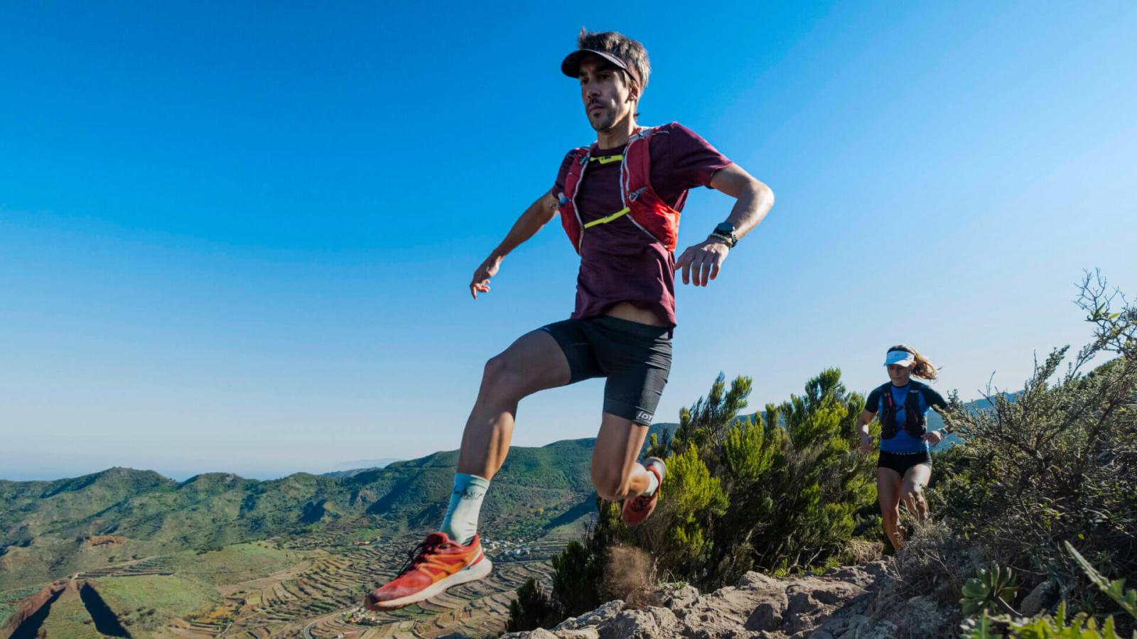 Personnes en train de courir sur un sentier rocheux dans un environnement naturel montagneux avec de la végétation et un ciel dégagé.