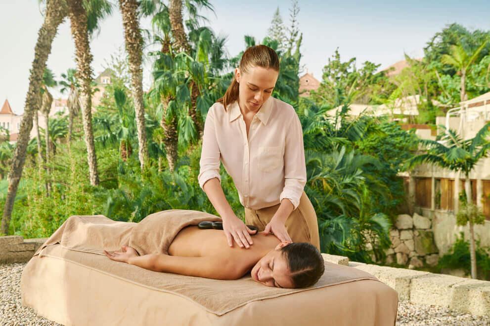 Thérapeute en train de réaliser un massage relaxant à l’air libre sur une table de massage, avec des palmiers et une végétation tropicale autour.