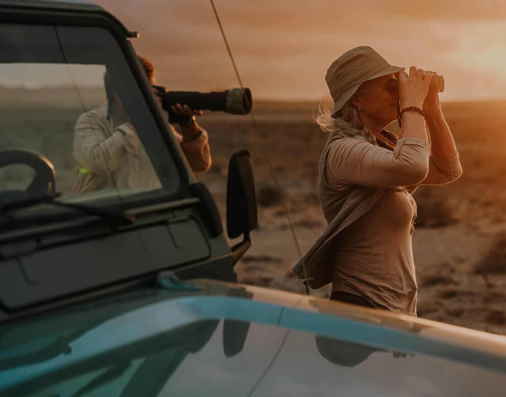 Personne observant les oiseaux avec des jumelles dans un paysage aride au coucher du soleil, près d'un 4x4.