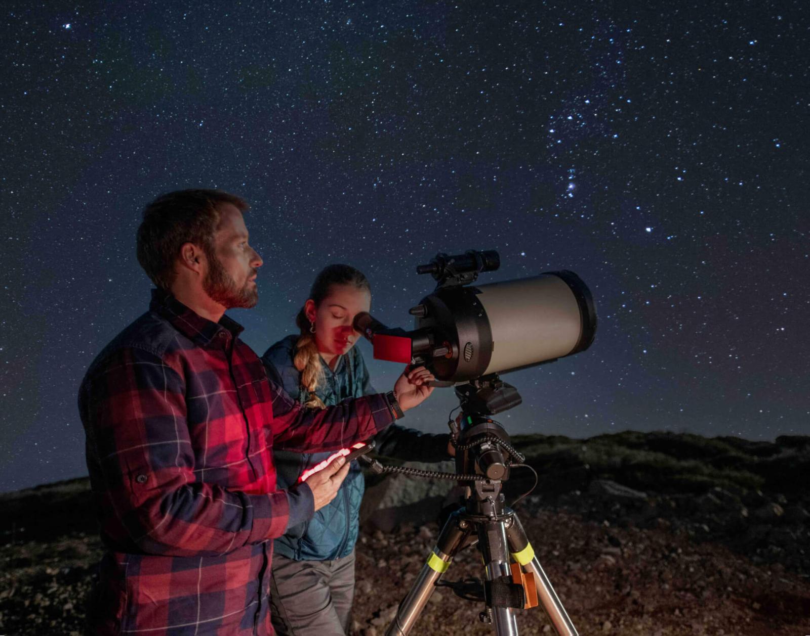 Deux personnes regardant au télescope sous un ciel nocturne étoilé, dans un paysage rocheux.