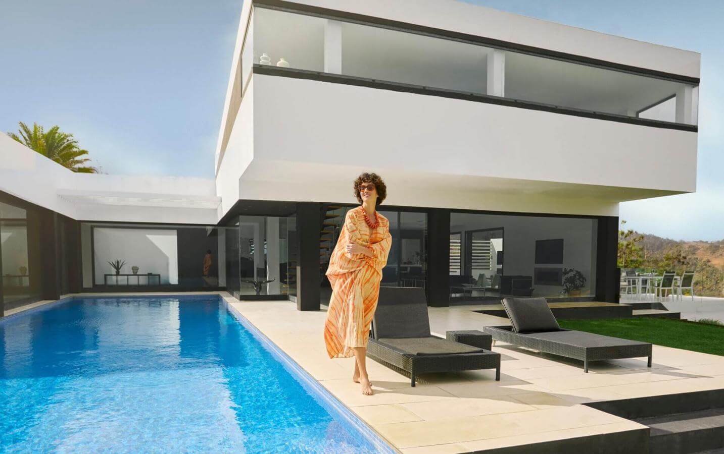 Personne se tenant au bord d’une piscine privée devant une villa contemporaine aux lignes épurées, avec des chaises longues et un ciel dégagé.