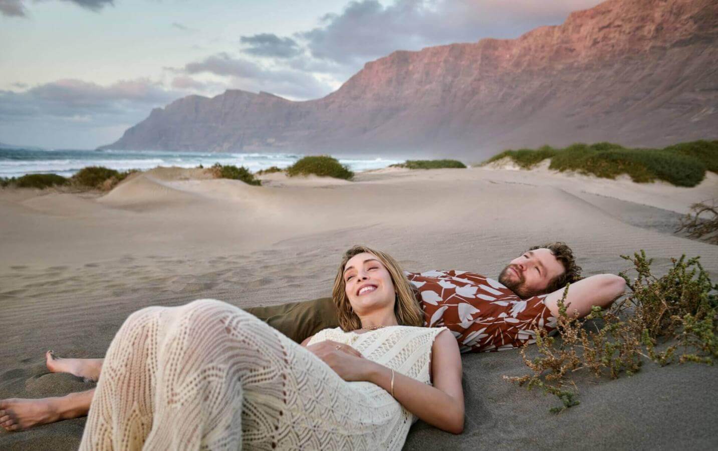 Deux personnes en train de se reposer sur le sable sur une plage sauvage, avec des montagnes en toile de fond, dans un paysage naturel et paisible.