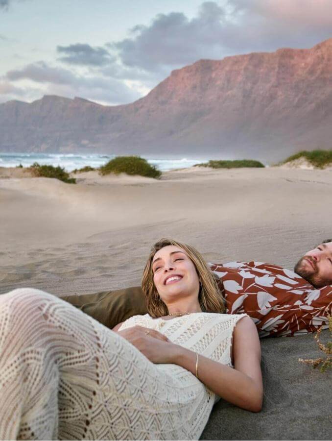 Deux personnes en train de se reposer sur le sable sur une plage sauvage, avec des montagnes en toile de fond, dans un paysage naturel et paisible.