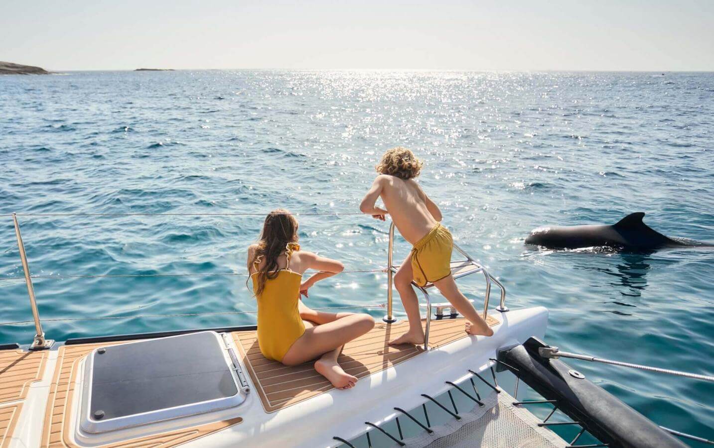 Deux personnes sur un voilier en train d’observer un dauphin dans des eaux cristallines au cours d’une sortie en mer par une journée ensoleillée.