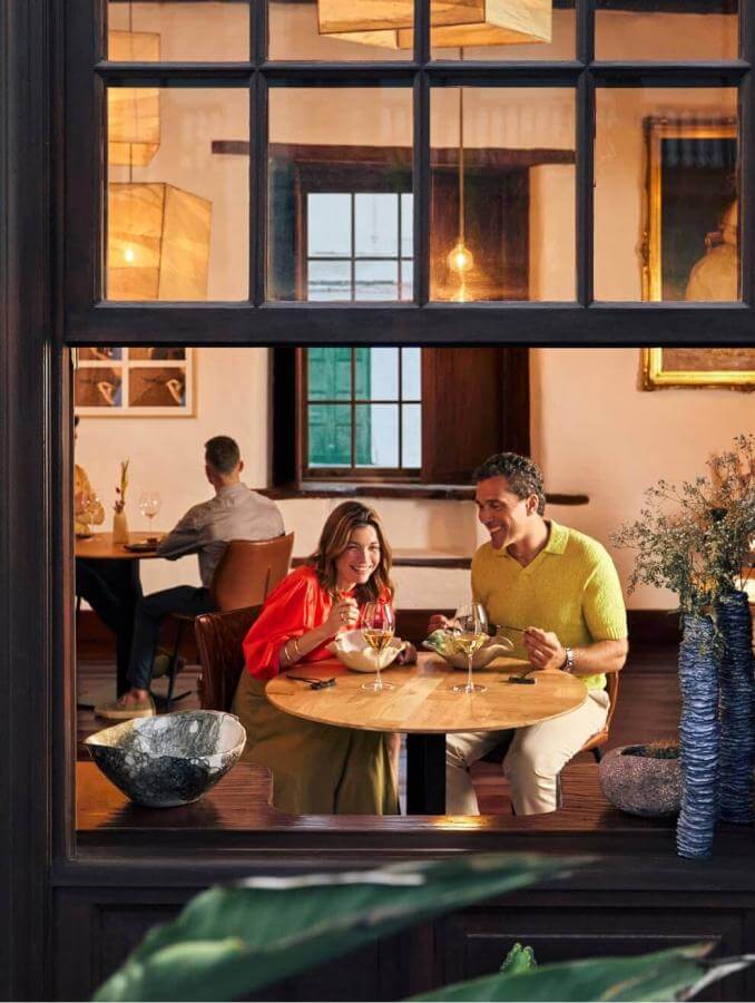 Vue à travers la fenêtre d’un couple en train de manger dans un restaurant chaleureux, avec une décoration traditionnelle et des détails naturels.